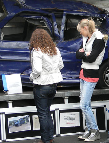zwei junge Damen schauen sich das ausgestellte Unfall-Fahrzeug mit den Beschreibungen zum Unfall an